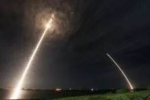Long-exposure photograph of a SpaceX Falcon 9 cargo rocket launching toward the International Space Station at night, with its returning booster tracing a second arc of light