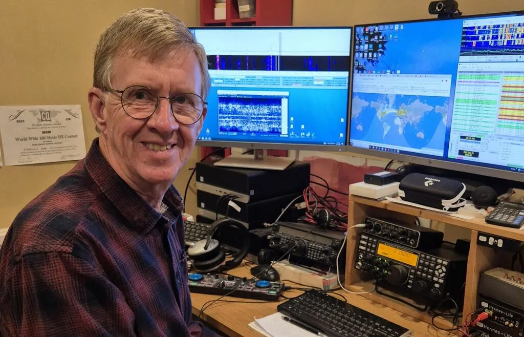 An amateur radio operator smiles at his contest station with two monitors showing logging and propagation software and HF transceivers on the desk.