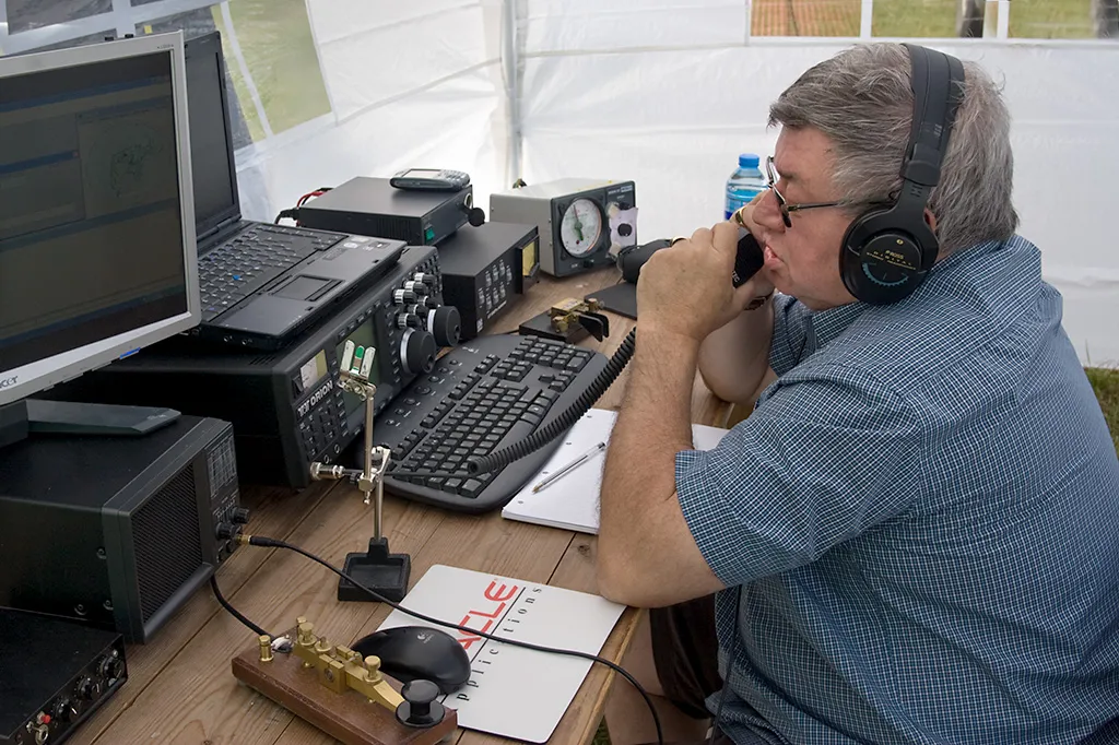 An amateur radio operator wearing headphones speaks into a microphone at a contest station with a transceiver, laptop logging computer, and Morse key on the desk.