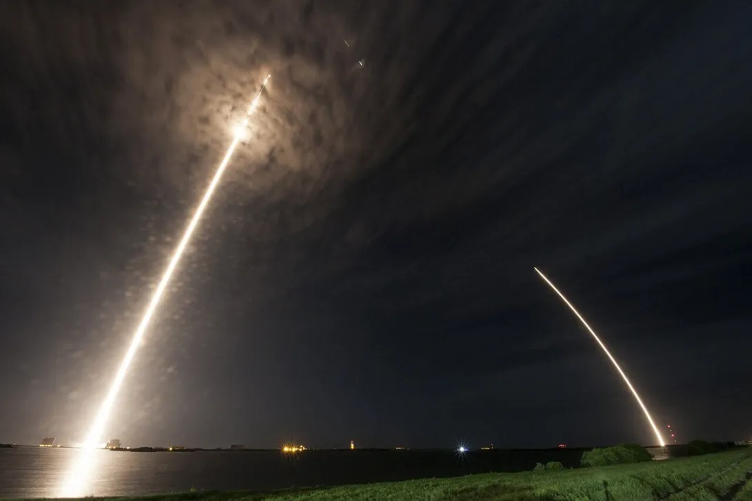 Long-exposure photograph of a SpaceX Falcon 9 cargo rocket launching toward the International Space Station at night, with its returning booster tracing a second arc of light